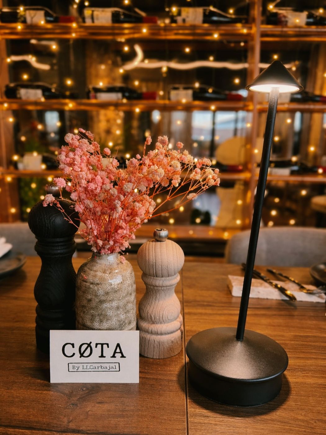 Mesa con lámpara y flores delante del botellero iluminado con guirnaldas