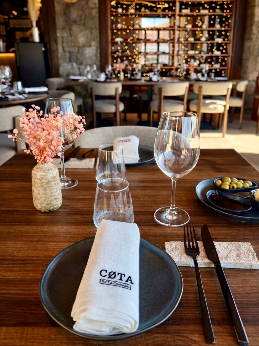 Mesa puesta en CØTA, con la servilleta bordada, copas y flores secas; al fondo, el comedor